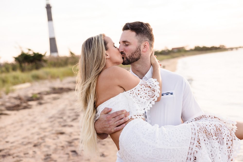 man holding woman in arms on beach with lighthouse in back