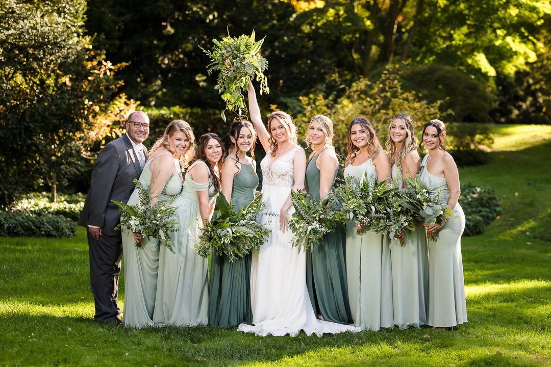 bridal party with bride holding up bouquet on grass at Bailey Arboretum