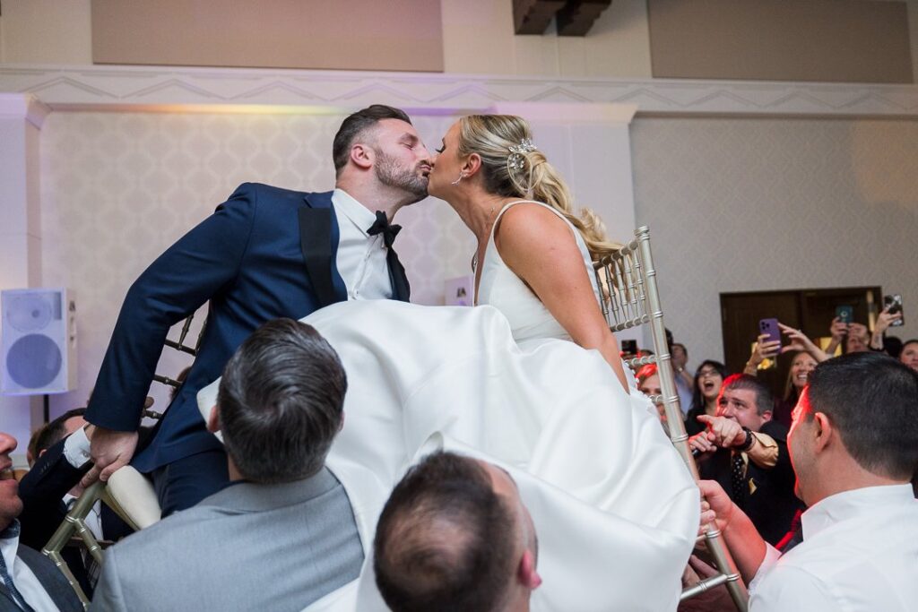 bride groom on chairs lifted by guests while kissing at Gatsby On The Ocean