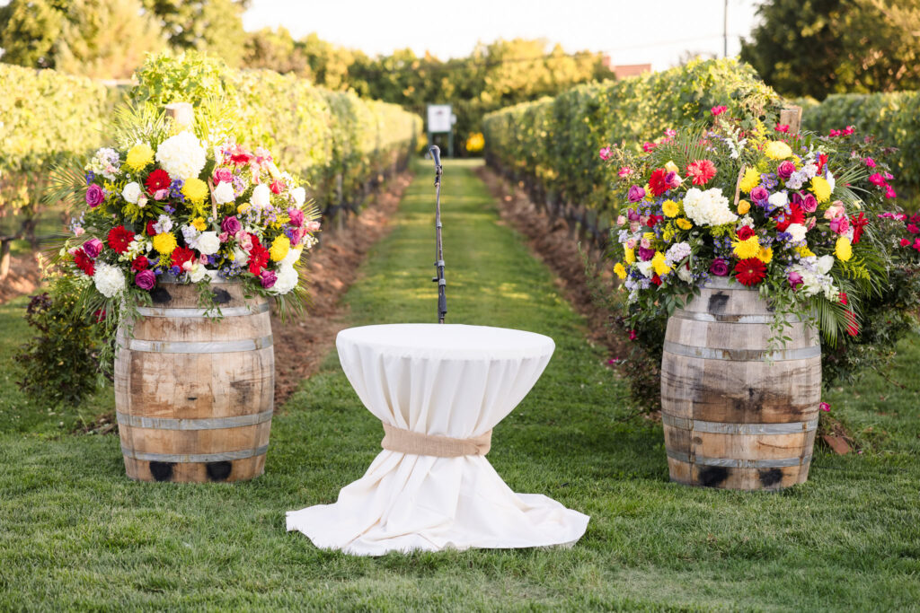 Outdoor vineyard wedding ceremony setup at Baiting Hollow Farm Vineyard, featuring a rustic aisle lined with grapevines, floral arrangements atop wine barrels, and a draped table with a microphone for vows.