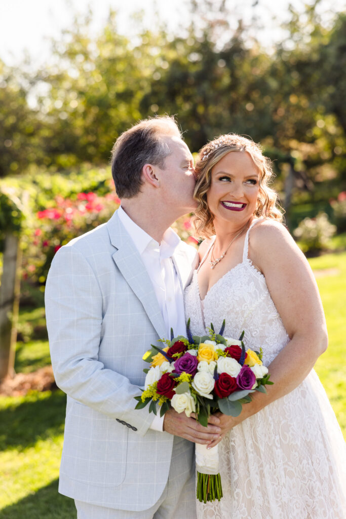 Joyful bride and groom share a tender moment among vineyard rows at Baiting Hollow Farm Vineyard, with the groom kissing the bride's cheek as she smiles, holding a vibrant bouquet of roses.