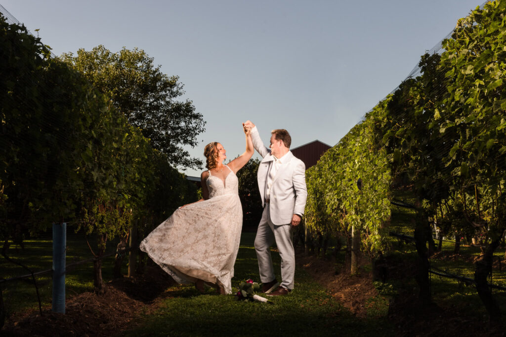 Bride and groom share a romantic twirl at golden hour between vineyard rows at Baiting Hollow Farm Vineyard, her lace gown catching the light as grapevines frame their joyful dance.