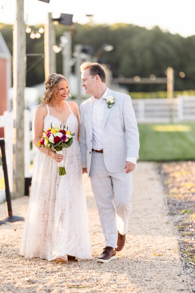 Newlyweds walk hand in hand along a sunlit path at Baiting Hollow Farm Vineyard, smiling at each other with joyful connection as string lights and rustic charm surround them.