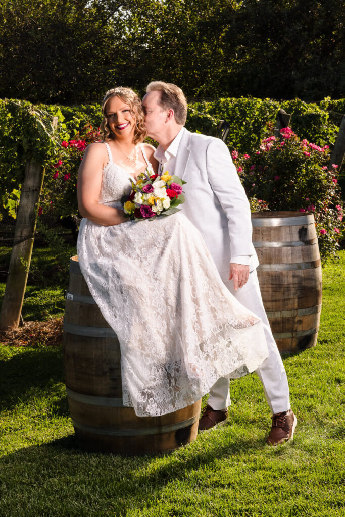 Bride sits playfully on a wine barrel with her groom by her side at Baiting Hollow Farm Vineyard, surrounded by blooming roses and lush vines, sharing a joyful kiss and vibrant bouquet.
