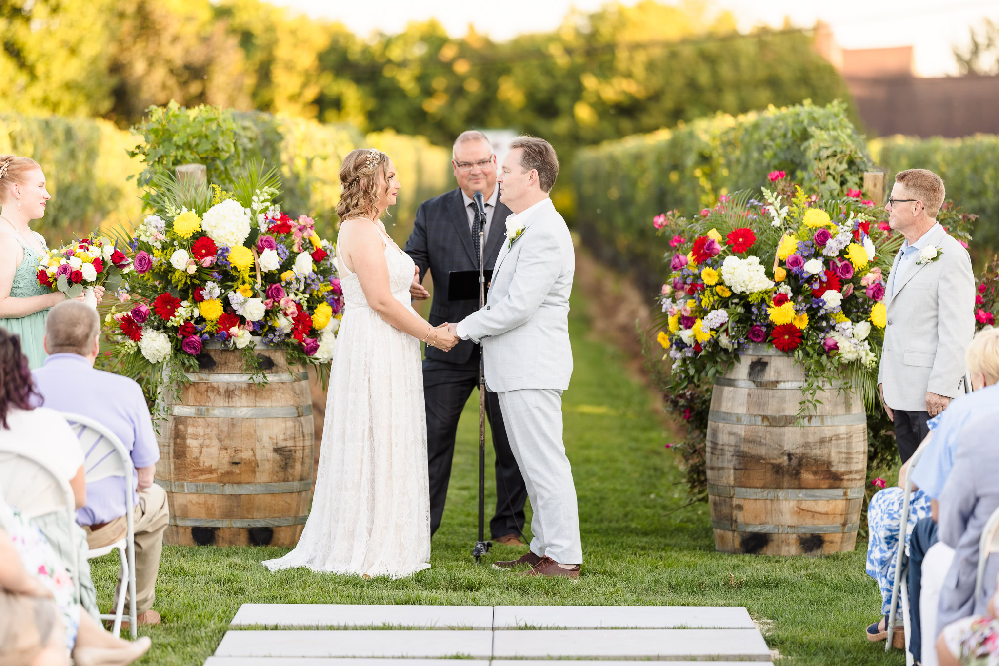 Bride and groom exchange vows during an outdoor vineyard ceremony at Baiting Hollow Farm Vineyard, framed by lush grapevines and vibrant floral arrangements atop rustic wine barrels.