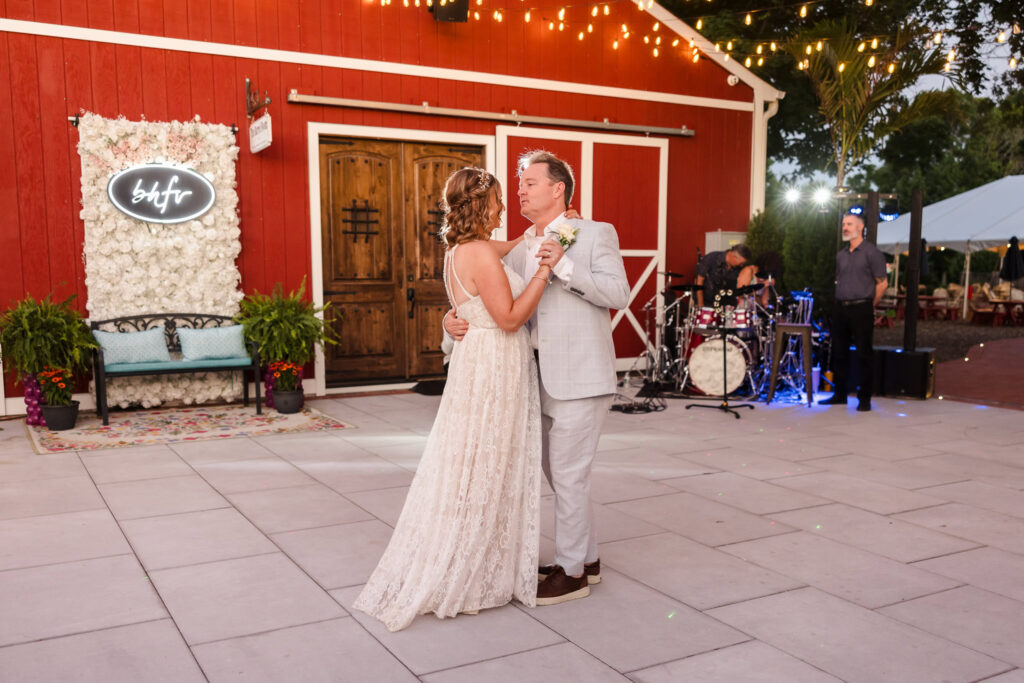 Bride and groom share their first dance under string lights beside the red barn at Baiting Hollow Farm Vineyard, surrounded by live music, soft evening light, and floral decor.