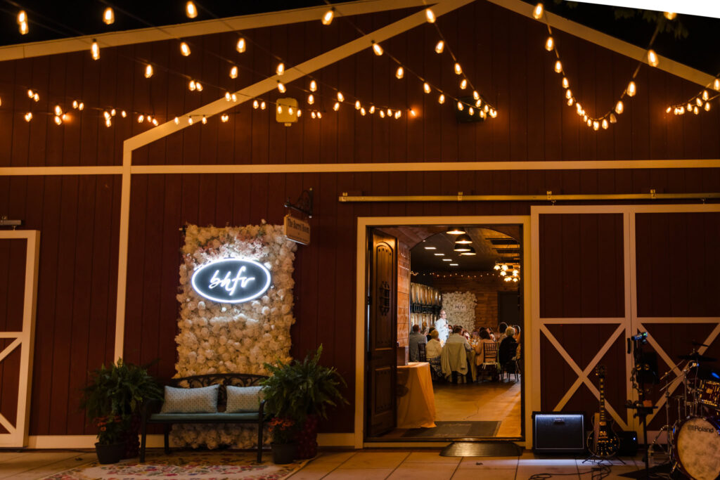 Evening reception at Baiting Hollow Farm Vineyard with glowing string lights, a floral photo backdrop with bhfv sign, and guests dining inside the warmly lit Barrel Room visible through open barn doors.