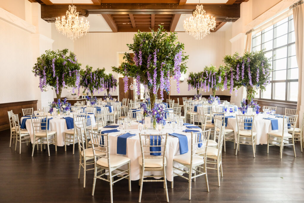 Ballroom at Gatsby on the Ocean set for a wedding with tall floral centerpieces, chandeliers, and round tables with silver chairs and navy accents.