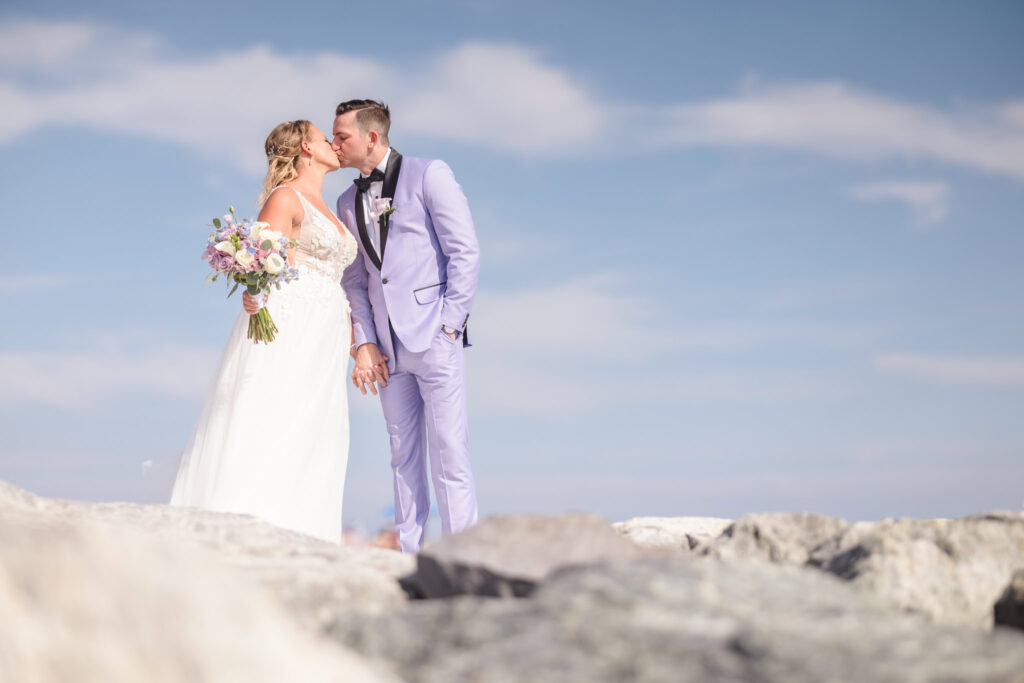 Groom and bride kissing while standing on rocks at the beach.