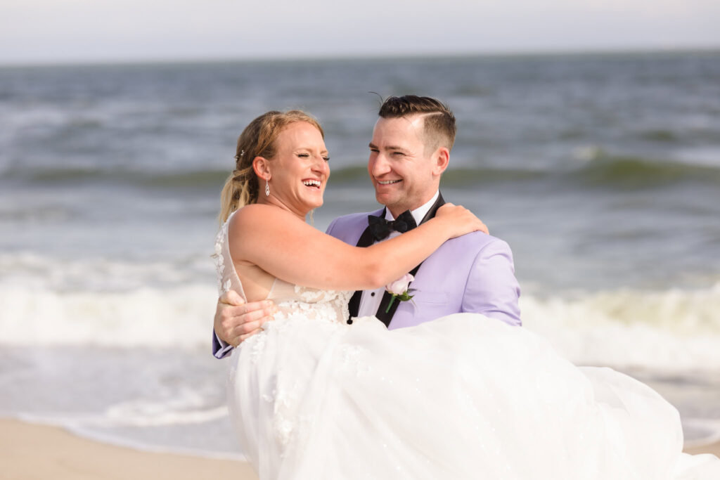 Groom carrying bride while they laugh on the beach.