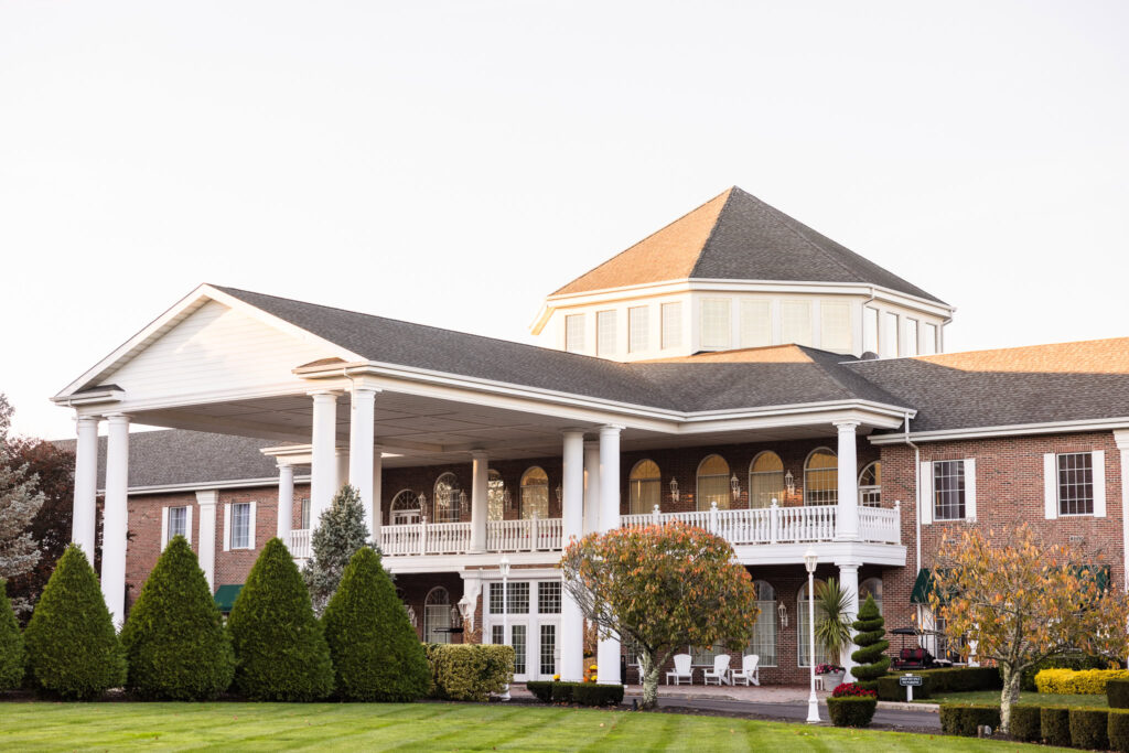 The grand exterior of East Wind Long Island, featuring a stately brick façade, white columned portico, manicured lawns, and a distinctive rotunda-style rooftop at golden hour.