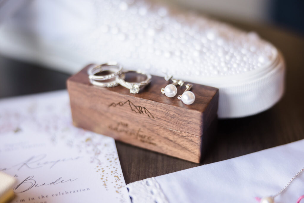 Wedding rings and earrings on custom engraved ring box next to white shoes and wedding invitation