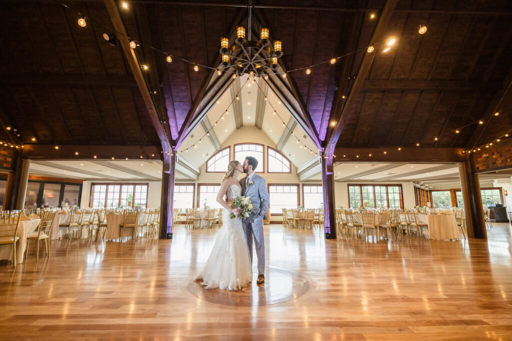 Bride and groom kissing beneath exposed wood beams and string lights inside The Riviera’s reception space.