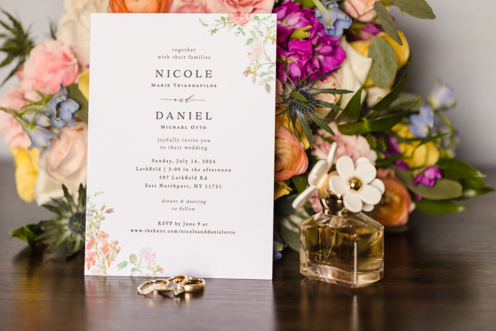 Bridal details including a bouquet, an invitation, rings, and perfume on a brown table.