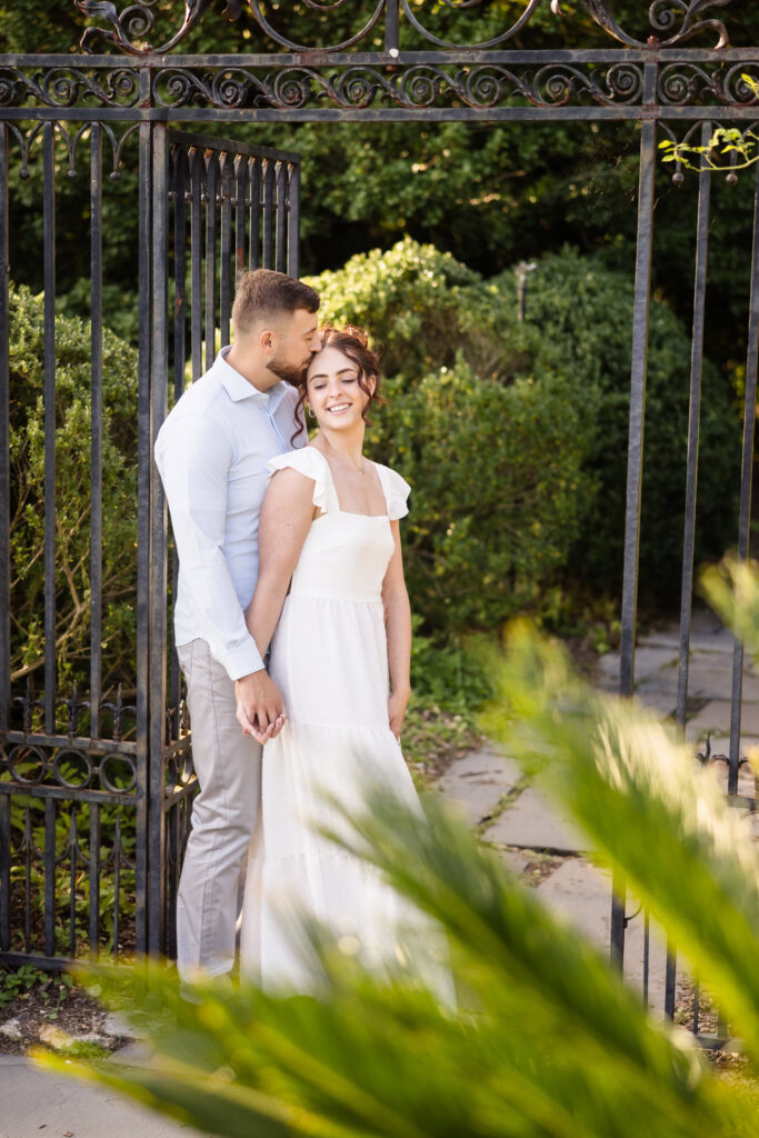 Man kissing woman on forehead at gate inside Planting Fields Arboretum engagement session.