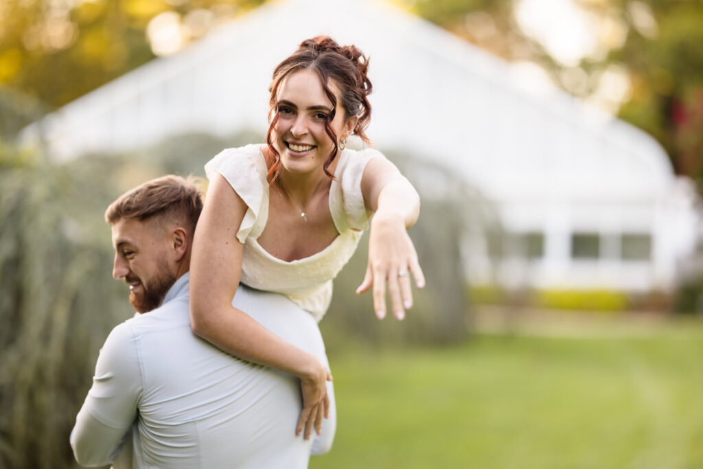 Woman on man's shoulder showing her engagement ring to the camera at Planting Fields Arboretum.