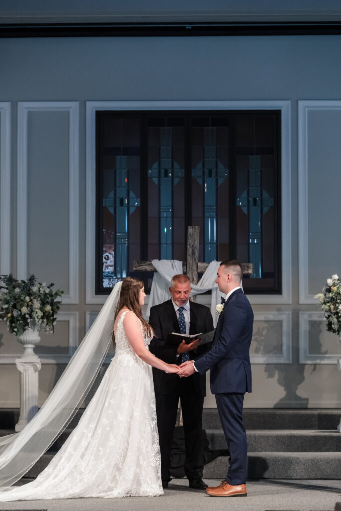 Bride and groom holding hands during the wedding ceremony with the pastor in the background.