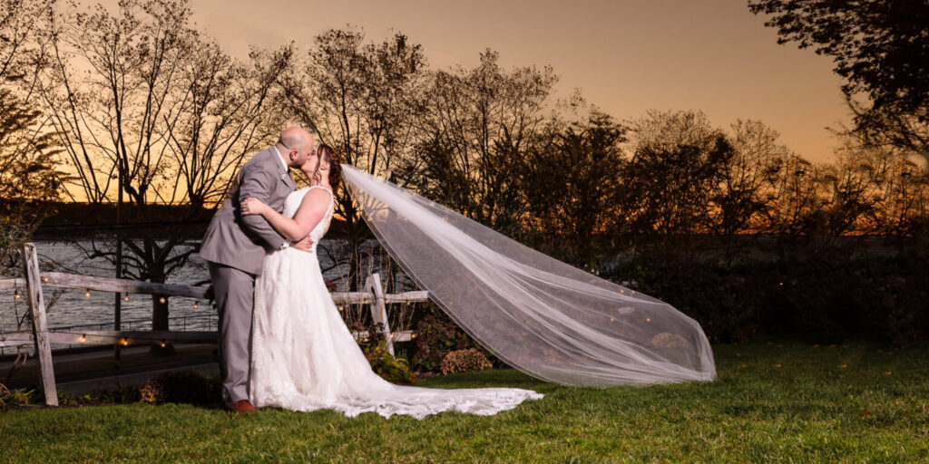Bride and groom kissing beside the water at sunset with the bride’s long veil flowing behind her at Sea Cliff Manor.