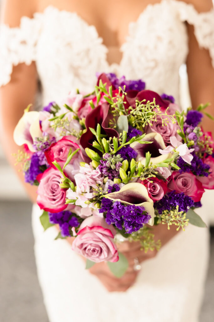 Close up of bride holding her bouquet. The bouquet has pink, purple, white, and green flowers.