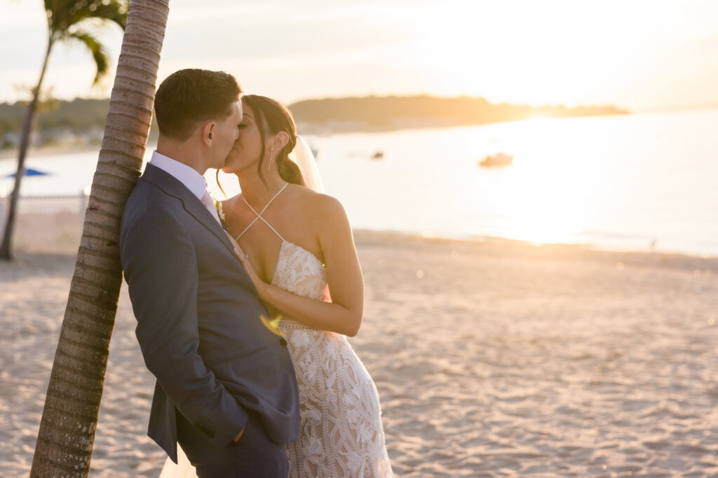 Bride and groom kissing on the beach while leaning on a palm tree Crescent Beach Club during sunset.