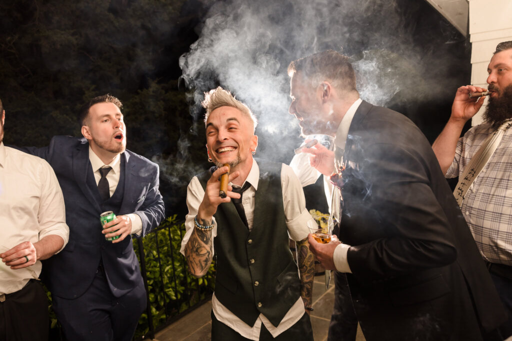 Groom smoking a cigar and laughing with friends at Bates House. 