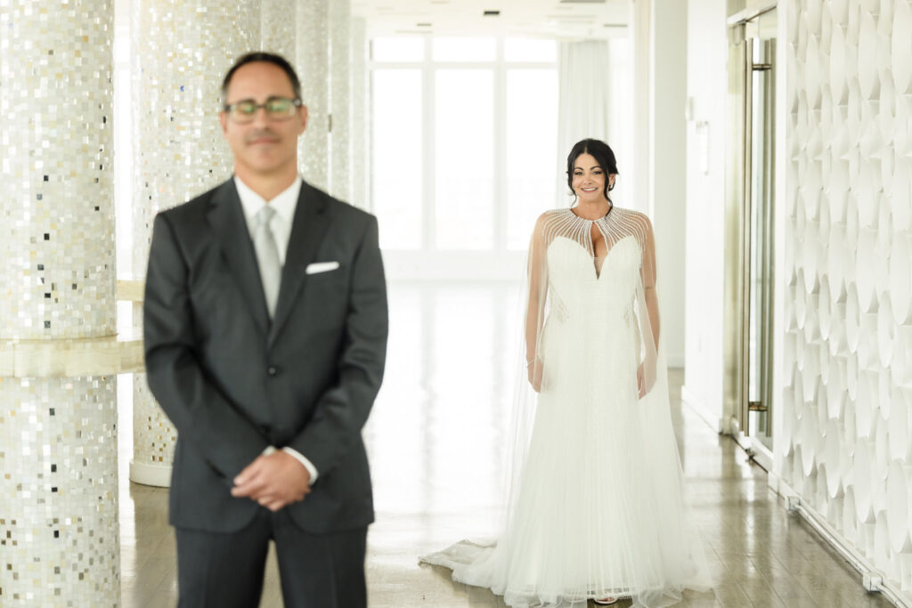 Bride in focus walking up to the groom before the first look on the rooftop bar of the Allegria Hotel in Long Beach. The groom is out of focus and has his eyes closed. The bride is smiling while looking at the groom. 