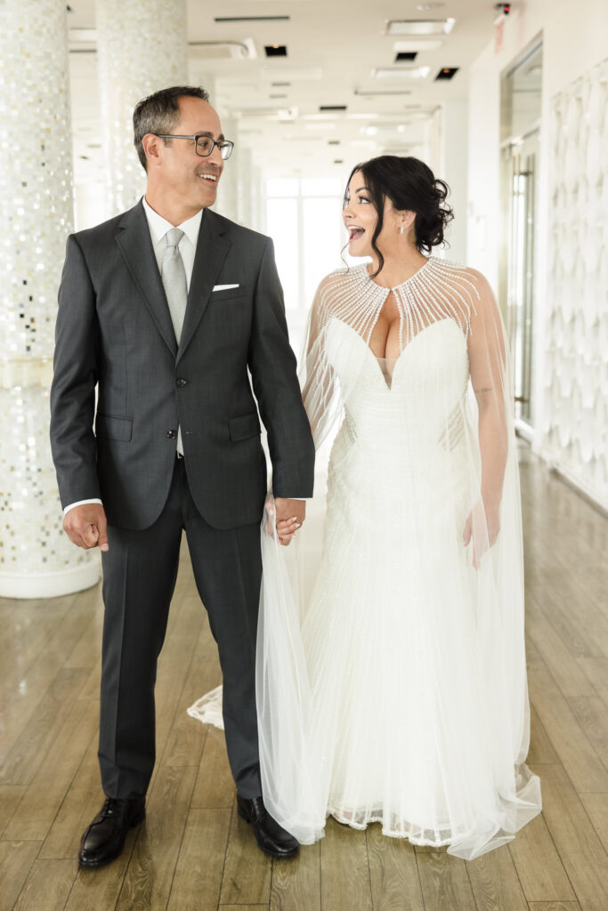 Bride and groom holding hands and seeing each other for the first time. They are on the rooftop of the Allegria hotel and both are smiling broadly. 