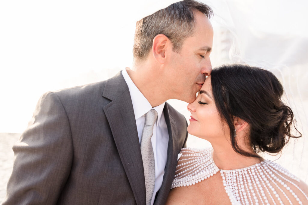 Groom kissing bride's forehead. Both of them have their eyes closed, and the bride is smiling. 