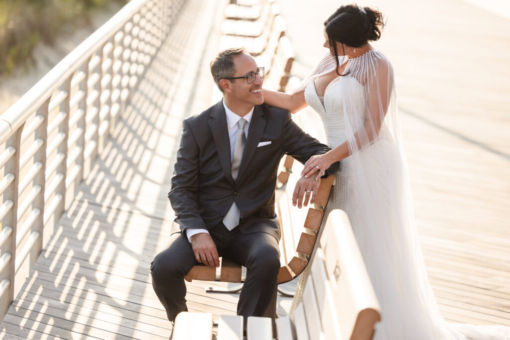 Bride standing next to the groom on the Long Beach boardwalk. The groom is sitting on a bench. 