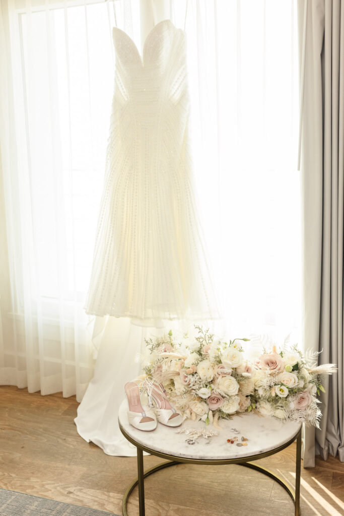 Wedding dress hanging in a window with shoes, bouquets, and jewelry on a marble table at Allegria Hotel.