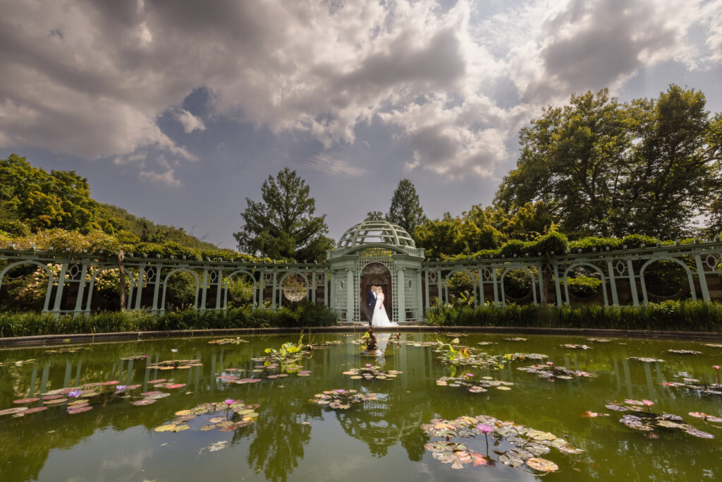 Bride and groom under and arch and in front of a pond at Old Westbury Gardens. The bride and groom are very small in the frame. 