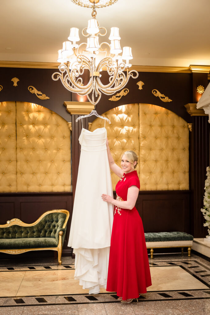 Bride in red dress for Chinese Tea Ceremony next to her Western dress hanging on a chandelier. 