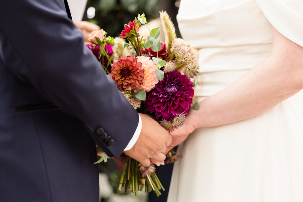 Close up of bride and groom's hands holding flowers during wedding ceremony at Sand Castle. 