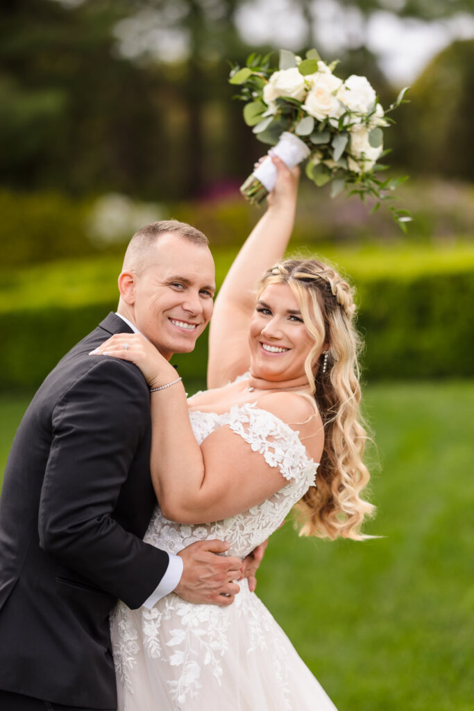 Bride and groom smiling at the camera. The bride is holding her white bouquet over her head.