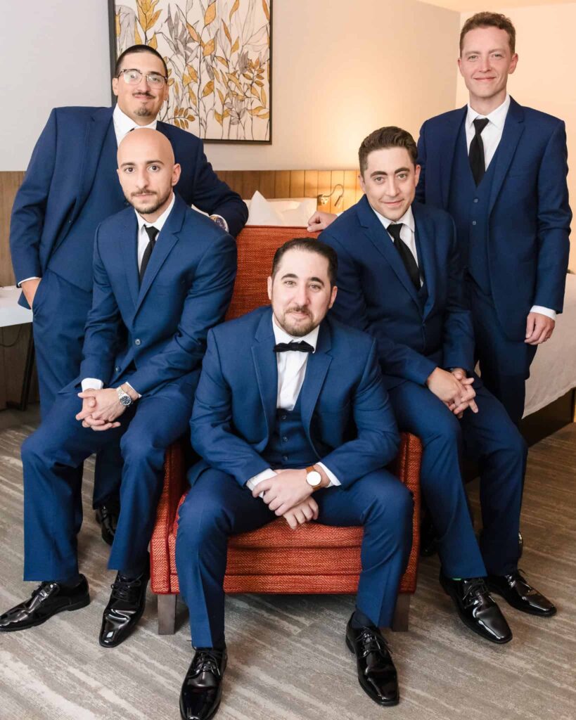 Groom and groomsmen in hotel room. Groom is sitting on a chair.