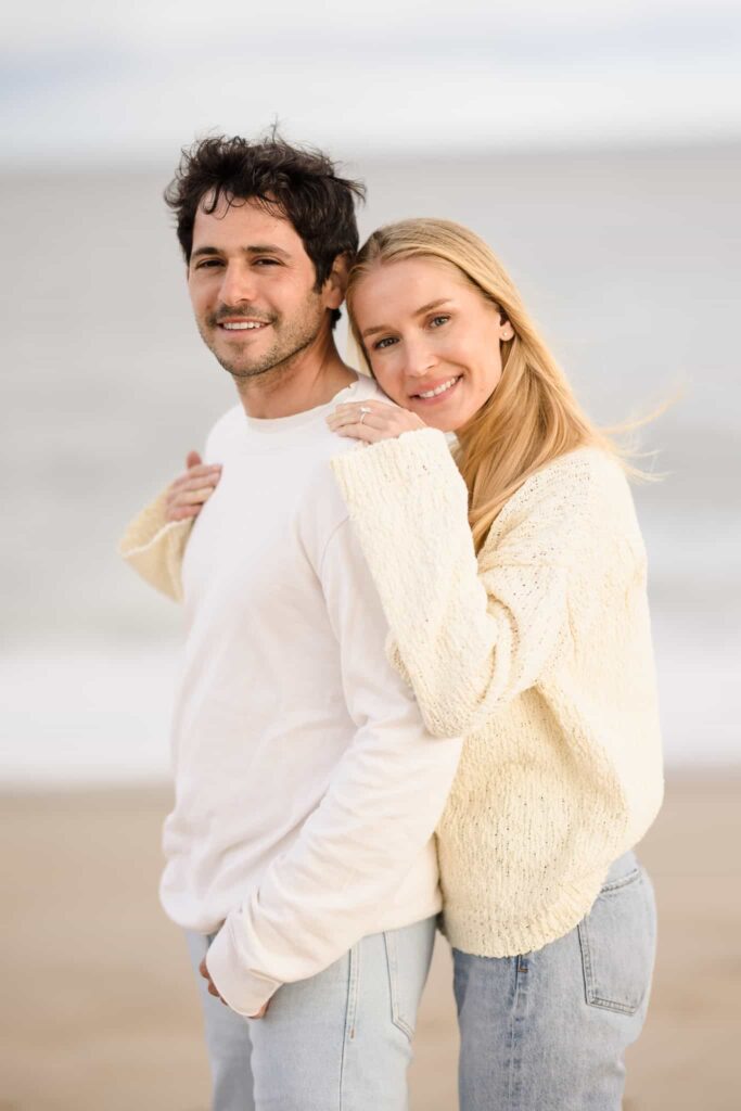 Woman hugging man from behind on the beach. Their engagement session outfits are white shirts with blue jeans. 