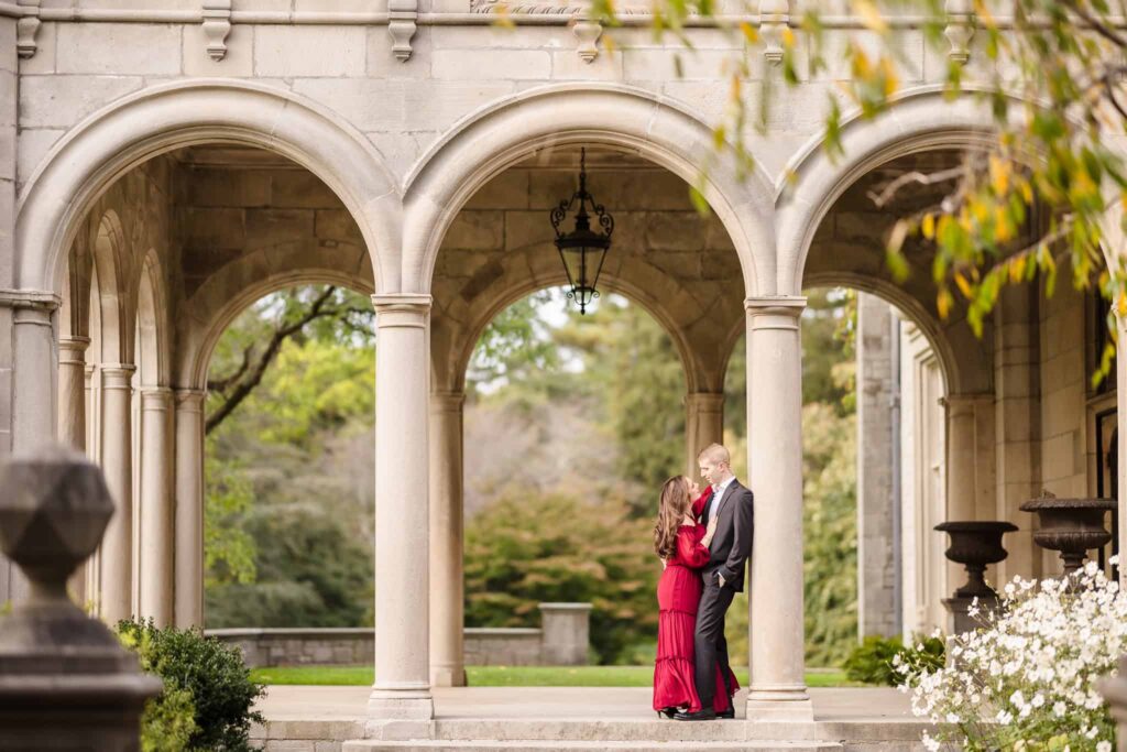 An engagement couple is kissing under arches at Planting Fields Arboretum. Their engagement session outfits include a burgundy dress and a dark suit. 