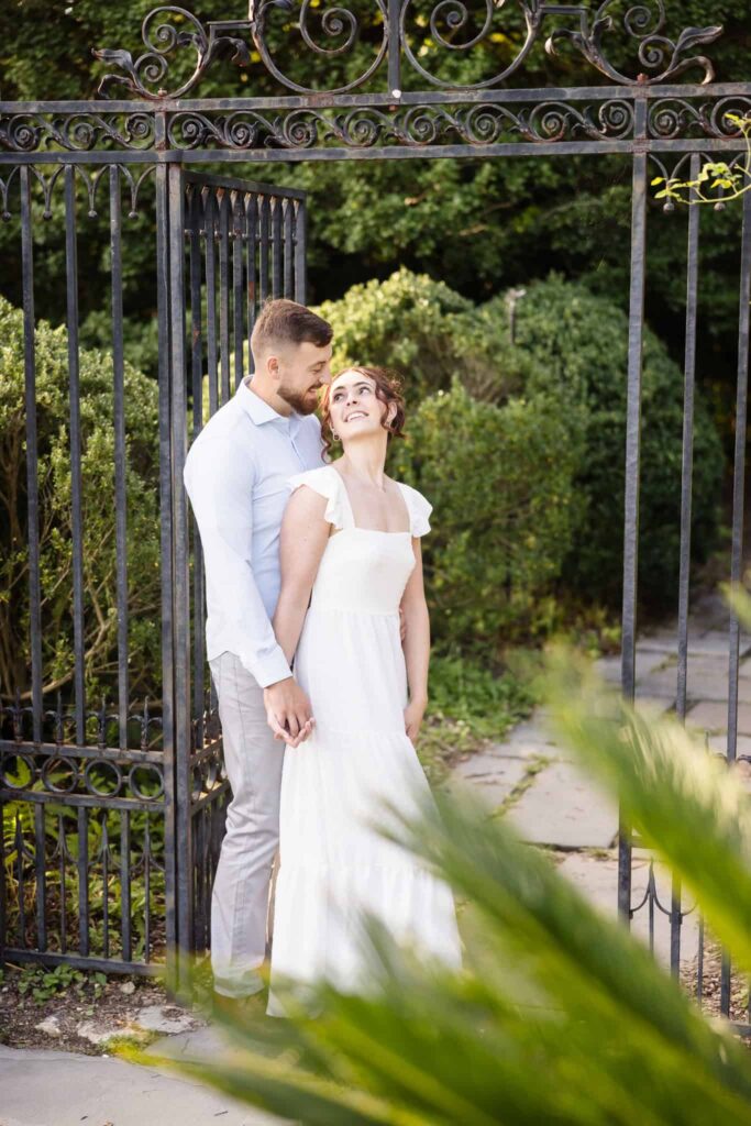 Couple at a gate smiling at each other. The woman is dressed in white and the man is dressed in blue. They are at Planting Fields Arboretum.