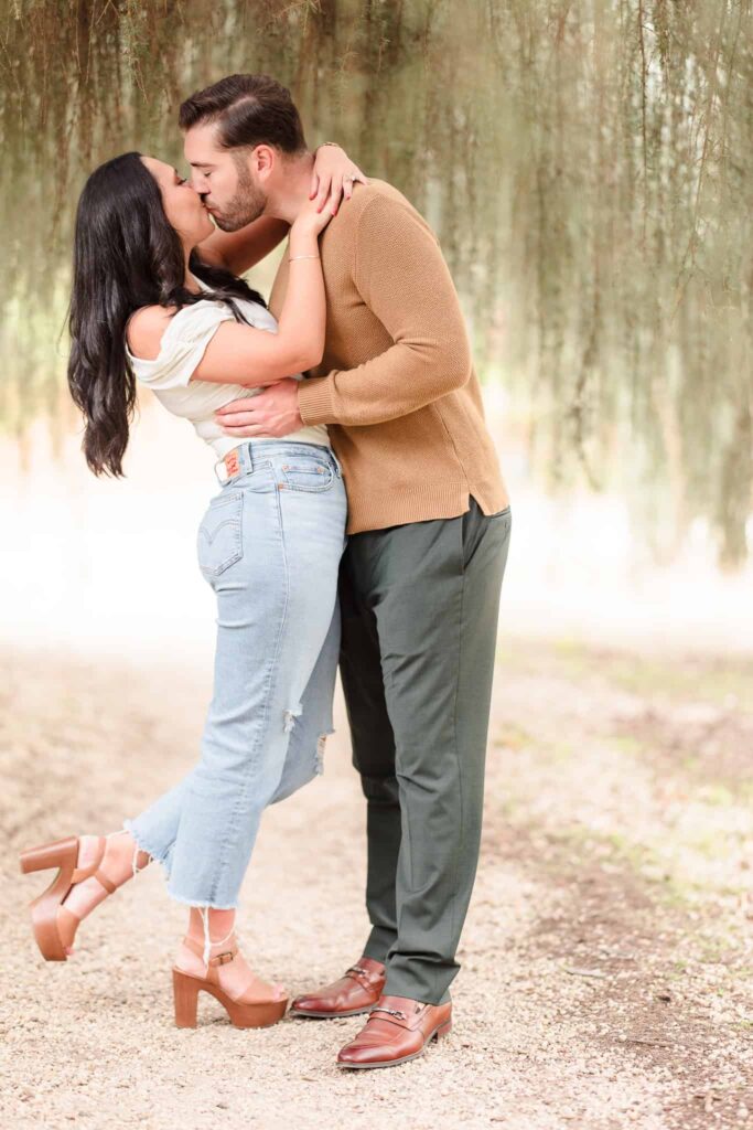 Man and woman in casual outfits at Planting Fields Arboretum. Their engagement session outfits include jeans with a white shirt. He is wearing a brown sweater and green pants. 