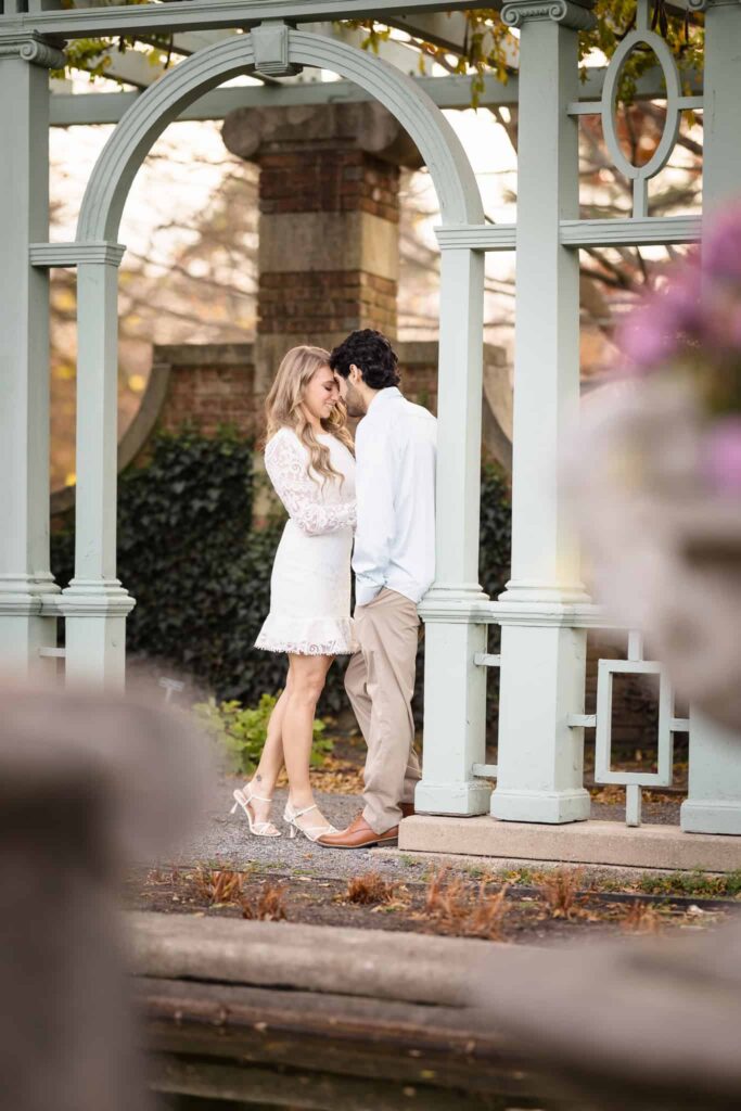 Man and woman under arch touching foreheads at Old Westbury Gardens. Their engagement session outfits include a white dress and he is wearing a blue shirt.