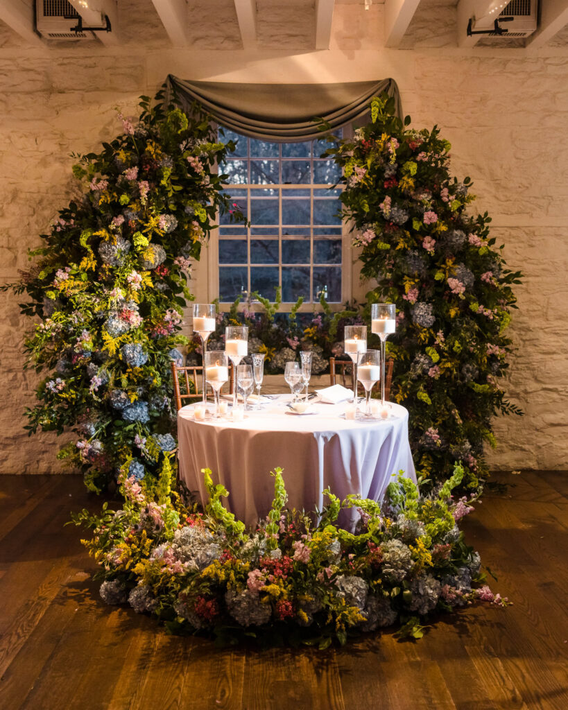 Sweetheart table at Stone Mill for wedding at New York Botanical Garden. There is greenery in front and behind the table.