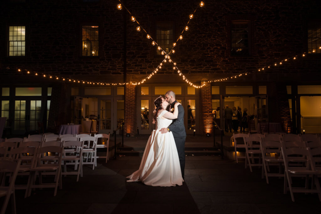 Bride and groom kissing under lights at night on patio at NYCB Stone Mill.