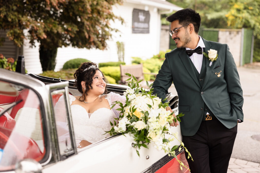 Groom leaning against a classic car with bride inside in front of WaterView at Port Jefferson. They are looking at each other and smiling.