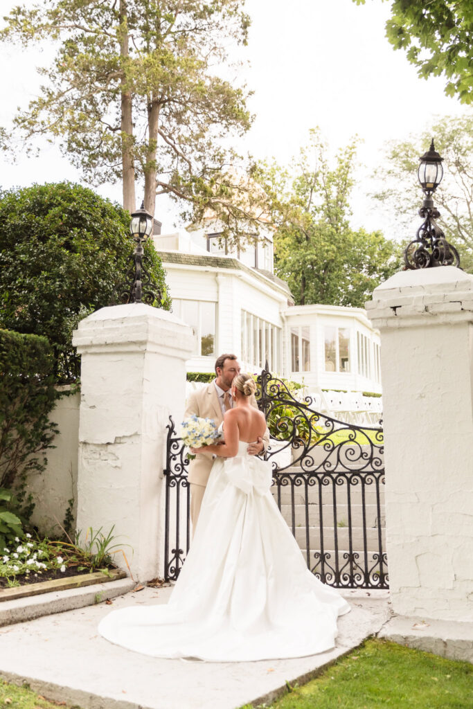 Bride and groom standing together between white brick pillars and ornate wrought-iron gates, with the manor visible behind them at Sea Cliff Manor.
