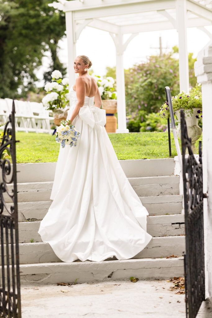 Bride standing on white garden steps and looking back toward the camera while holding a blue-and-white bouquet, with a white arbor behind her at Sea Cliff Manor.