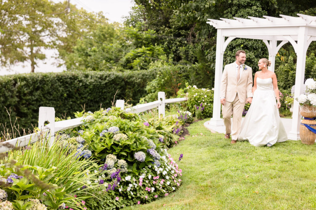 Bride and groom holding hands as they walk through the hydrangea-filled garden beneath a white pergola at Sea Cliff Manor on Long Island.