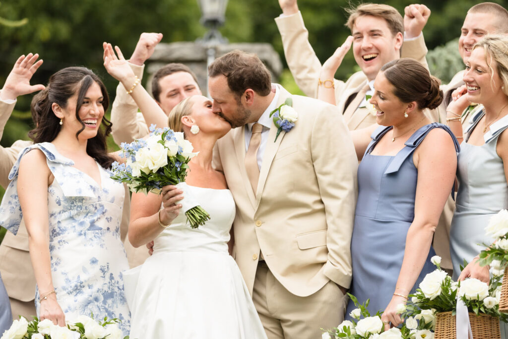 Bride and groom kissing in the center of their wedding party as bridesmaids and groomsmen gather around them with raised arms at Sea Cliff Manor.
