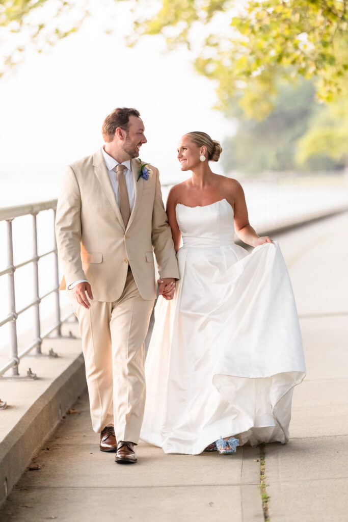 Bride and groom holding hands and looking at each other as they walk along the waterfront at Sea Cliff Manor. The bride lifts the skirt of her wedding dress as they walk.