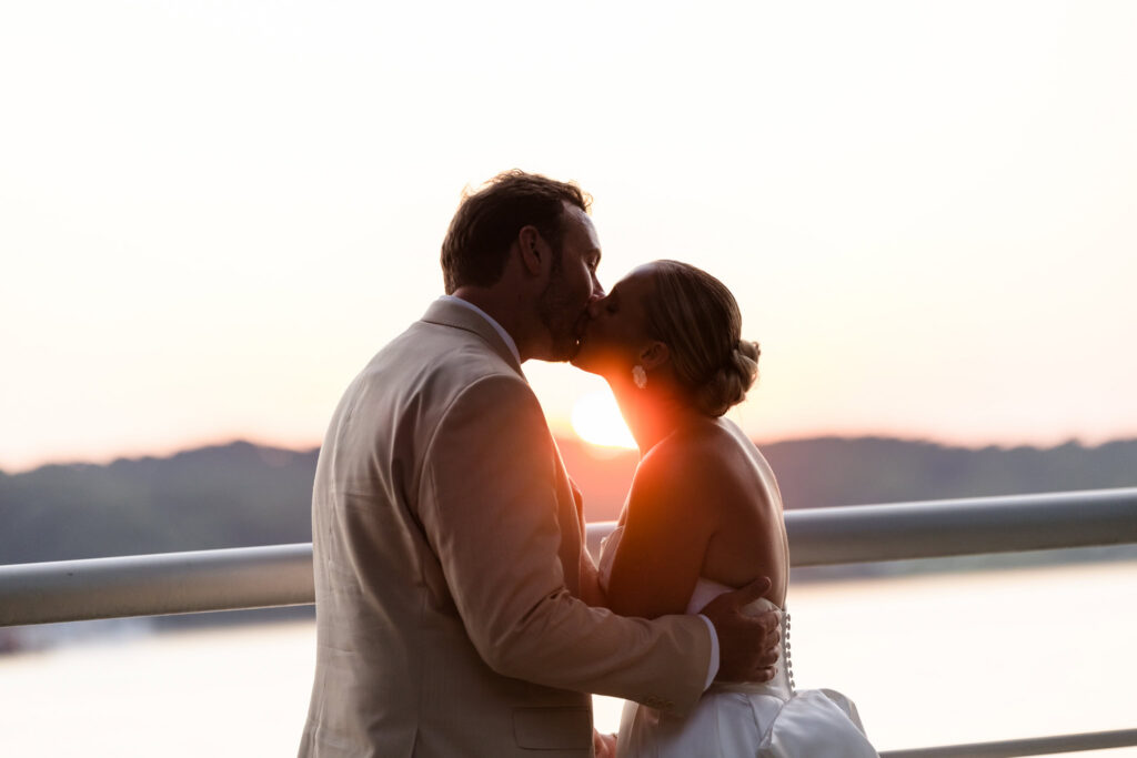 Bride and groom kissing beside the waterfront railing at sunset, with the sun between them and Hempstead Harbor behind them at Sea Cliff Manor.