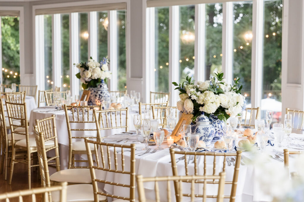 Wedding reception tables with white linens, gold Chiavari chairs, and blue-and-white floral centerpieces arranged beside large windows at Sea Cliff Manor.